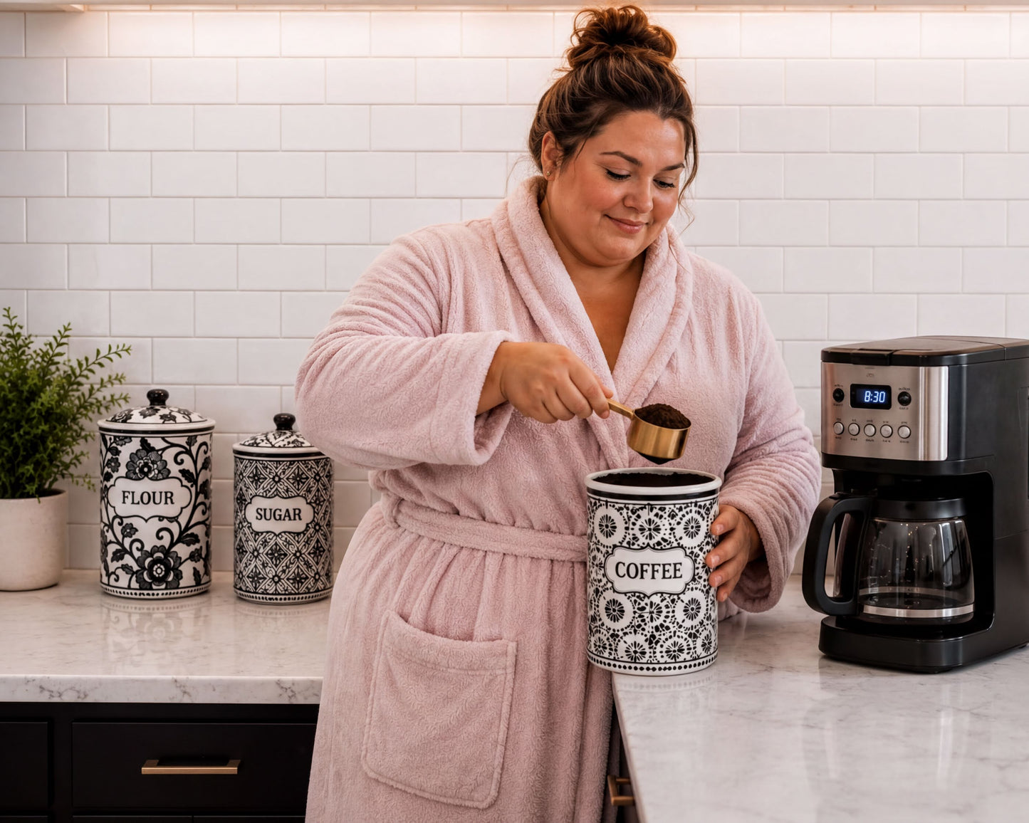 A woman in a pink robe is pouring coffee grounds into a coffee maker on a kitchen counter.